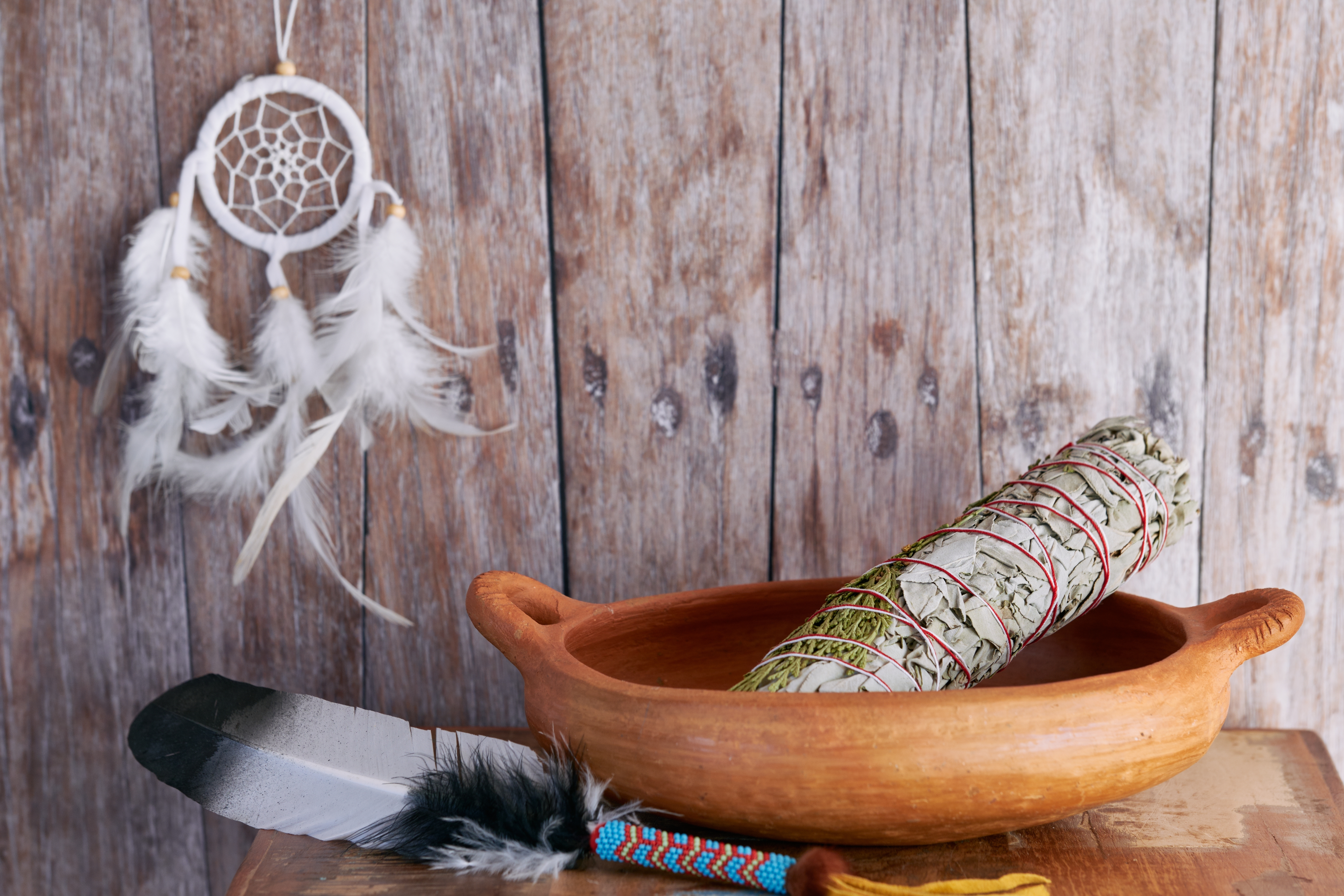 A smudge stick (bundle of dried white sage and juniper) in a clay bowl with a decorative feather to spread the smoke and a dreamcatcher in the background | Source: Shutterstock